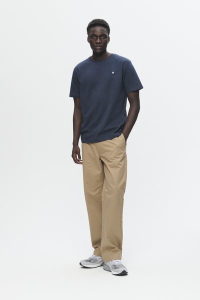 T-shirt col rond marine avec un petit logo, associé à un pantalon beige clair. Modèle portant des chaussures de sport grises avec des accents blancs.
