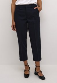 Pantalon bleu marine ajusté avec une silhouette raccourcie, présentant une texture lisse, deux poches avant et un devant plat. Stylé avec des chaussures à talons noires.