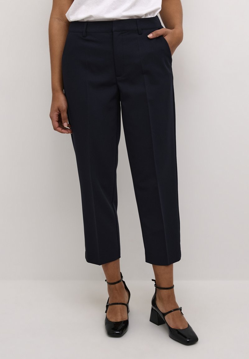 Pantalon bleu marine ajusté avec une silhouette raccourcie, présentant une texture lisse, deux poches avant et un devant plat. Stylé avec des chaussures à talons noires.