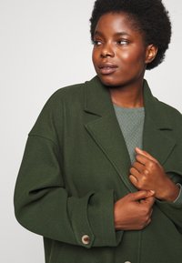 Green wool-blend coat with a notched collar, oversized fit, and buttoned cuff details, layered over a textured green sweater.