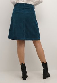 Teal corduroy skirt with a fitted waist, knee-length hem, and back pockets; textured fabric and straight silhouette. Black heeled ankle boots.