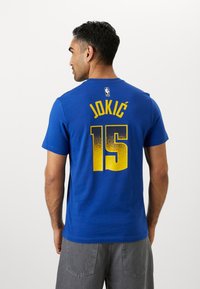 Zils kokvilnas T-krekls ar dzeltenu drukātu tekstu "JOKIĆ" un numuru "15" uz muguras, ar NBA logo etiķeti uz kakla.