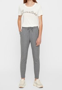 Vit t-shirt med texten "l'aventure" i svart kursiv, tillsammans med grå träningsbyxor med elastiskt midjeband och sidofickor. Beige sneakers.