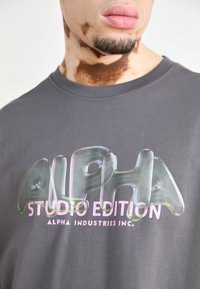 Pelēka kokvilnas T-krekls ar lielu, spīdīgu, 3D teksta dizainu iridescentās krāsās. Teksts ietver "ALPHA STUDIO EDITION" biezā, mūsdienīgā fontā.