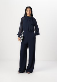 Mørkeblå jumpsuit med lange, gjennomsiktige ermer og høy hals. Vid bendesign, teksturert stoff og en stilren, sølvfarget clutch.