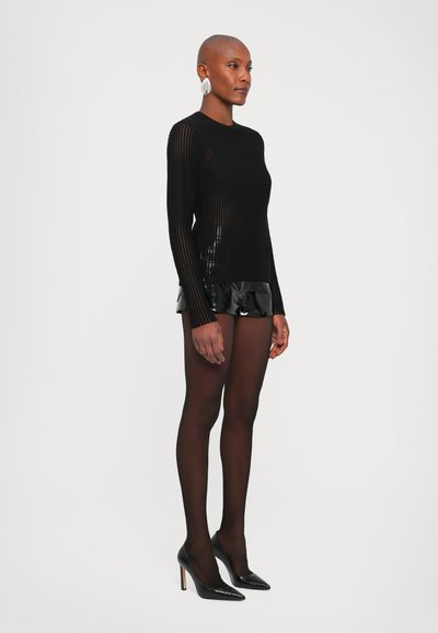 Black ribbed long-sleeve top paired with shiny black patent shorts. Model wears sheer black tights and pointed black high heels. Silver earrings.