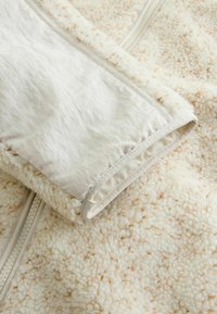 Fleece bunda s textúrovaným krémovým povrchom, ktorá má hladký biely vnútorný podšívku a zips. Povrch má jemný škvrnitý vzor.