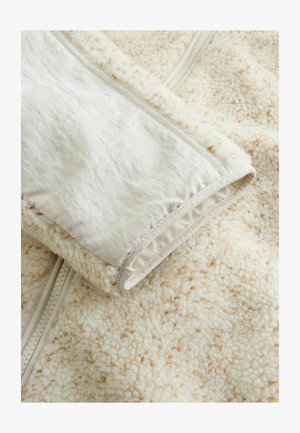 Fleece-takki, jossa on teksturoitu kerma-sisus ja sileä valkoinen vuori sekä vetoketju. Pintaa koristaa hillitty laikkuinen kuvio.