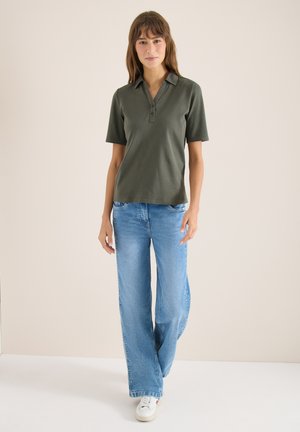 Femme portant un polo à manches courtes vert olive, un jean large bleu clair et des baskets blanches, debout devant un fond uni.