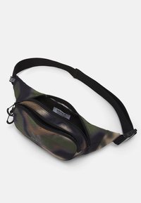 Riñonera de tela con patrón de camuflaje oscuro, con un compartimento principal con cremallera y correa ajustable negra. Presenta un logo de adidas en la parte frontal.