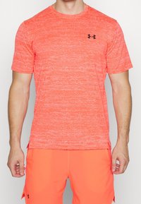 Kortärmad atletisk tröja i stark korallfärg med en strukturerad yta. Har en svart logotyp på bröstet och sidofickor för ventilation.