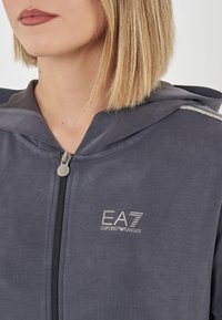 Felpa grigia con zip e cappuccio, con un logo EA7 lucido sul petto e decorazioni sulle spalle. Tessuto morbido con una finitura liscia.