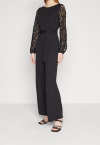 Svart jumpsuit med långa spetsärmar, figursydd midja knuten med ett bälte, bred ben-design, och matchad med svarta remsandaler.