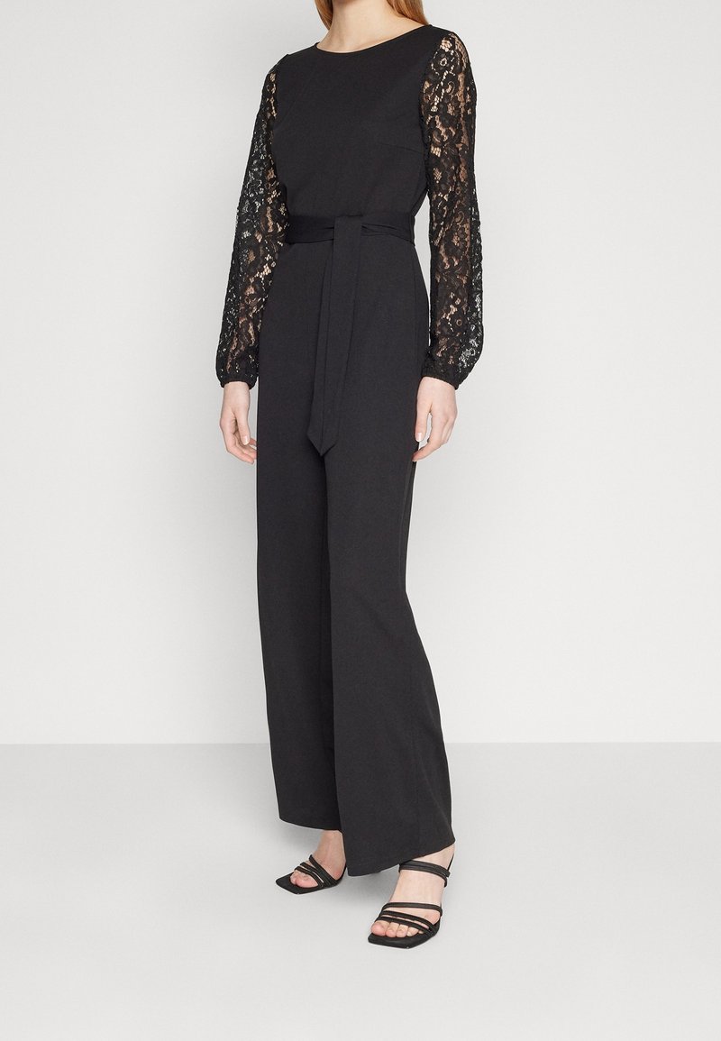 Svart jumpsuit med långa spetsärmar, figursydd midja knuten med ett bälte, bred ben-design, och matchad med svarta remsandaler.