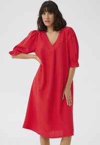 Robe rouge avec un décolleté en V, des manches bouffantes et une coupe midi. Fabriquée en tissu léger avec un motif texturé subtil.