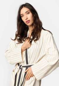 Bijela prugasta haljina s dugim rukavima i crnim vezanjem. Sadrži bočne džepove i mekan, strukturiran materijal prikladan za odjeću za opuštanje.