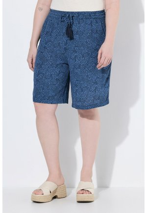 Person trägt blaue gemusterte knielange Shorts mit Seitentaschen und beige Plateausandalen und steht an einer weißen Wand.