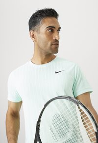 Kerge sinises Nike särkis mees hoiab tennisereketit ja vaatab kõrvale lihtsa taustaga.