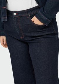 Temno modre kavbojke iz jeansa z rdečimi šivi, z žepom spredaj in zapiranjem na gumb v pasu.