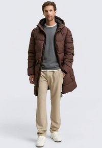 Braune, gesteppte Steppjacke mit Kapuze, durchgehendem Reißverschluss und Seitentaschen, darüber ein grauer Pullover und cremefarbene Hosen, kombiniert mit weißen Turnschuhen.