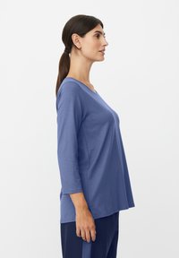 Niebieska bluzka z długim rękawem, z dekoltem w serek i bocznymi rozcięciami. Miękka, lekka tkanina o luźnym kroju. Połączona z ciemnymi spodniami.