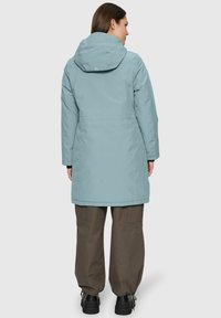 Vrouw in een lichtblauwe hoodie-parka, bruine cargobroek en zwarte laarzen, staande met haar rug naar de camera tegen een effen achtergrond.