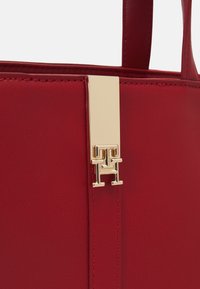 Gros plan d'un sac en cuir rouge avec une sangle verticale et une boucle en métal doré présentant un logo embossé "TH".