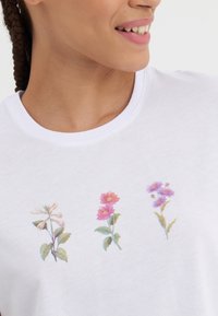 Camiseta blanca con tres flores bordadas: una flor blanca a la izquierda, una flor rosa en el centro y una flor morada a la derecha.