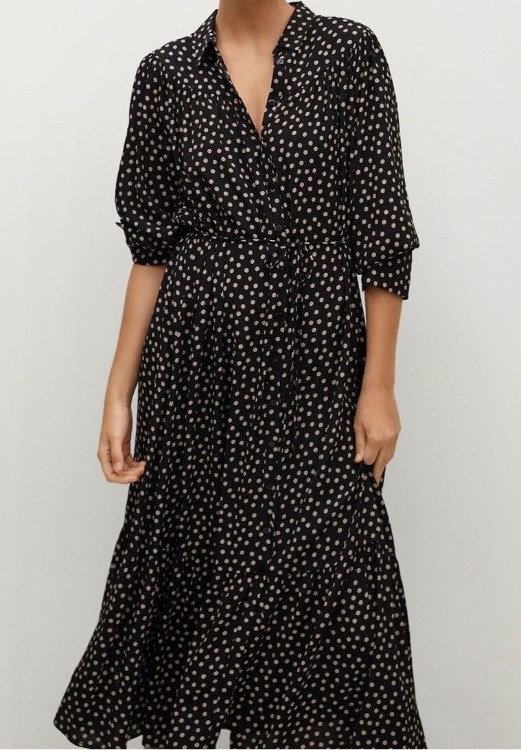 Femme portant une robe midi noire à pois beige, boutonnée sur le devant, manches trois-quarts et ceinture nouée à la taille.