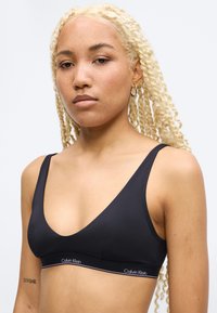 Γυναίκα με μακριά ξανθά πλεγμένα μαλλιά φοράει μαύρο bralette Calvin Klein και μικρούς κρίκους σκουλαρίκια, κοιτάζει μπροστά με ουδέτερη έκφραση.