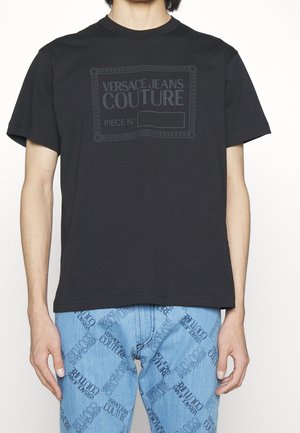 Camiseta de algodón negra con un estampado de logo en relieve con un borde de cuerda y el texto "VERSACE JEANS COUTURE". Diseño de manga corta y cuello redondo.