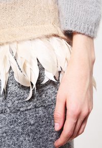 Beige gestricktes Kleidungsstück mit einer weichen Textur, verziert mit einem Federbesatz in sanften Tönen. Der graue Pullover darunter hat eine grobe, filzartige Textur.