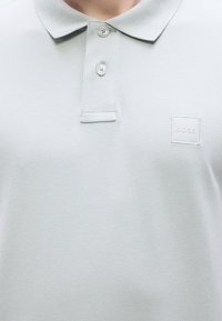 Ανοιχτό γκρι πόλο μπλούζα κατασκευασμένη από υφασμάτινη ύλη με υφή, με γιακά, δύο κουμπιά και λευκό τετράγωνο λογότυπο με τη λέξη "BOSS" στο μπροστινό μέρος.