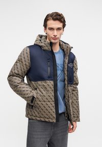 Jovem vestido com uma jaqueta beje e azul-marinho com padrão e capuz, uma t-shirt gráfica azul e jeans cinza-escuro, de pé contra um fundo simples.