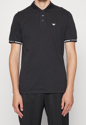 Uomo che indossa una polo nera Emporio Armani con tre bottoni e logo bianco sul petto e sui polsini delle maniche.