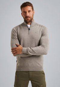 Šedá mikina na zip s dlouhými rukávy, s vysokým límcem a jemným pleteným vzorem. Nositá přes bílou košili, v kombinaci s olivově zelenými kalhotami.
