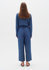Denim kombinezons vidējā zila krāsā ar garām piedurknēm, elastīgu jostasvietu, platu bikses kāju un aizmugurējo kabatu. Gluda tekstūra ar klasisku šuvju rakstu.