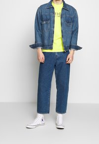 Chaqueta de mezclilla con un lavado claro, dos bolsillos en el pecho, combinada con una camiseta amarilla brillante y jeans de pierna recta azules; zapatillas altas blancas.