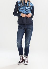 En person som bär en blå camouflagemönstrad kvartslång jacka, slitna uppvikta jeans och vinröda högskurna sneakers står med händerna knäppta.