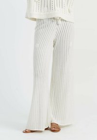 Pantaloni ampia in maglia bianca con strisce verticali e dettagli testurizzati, abbinati a sandali marroni con cinturini. Tessuto leggero.