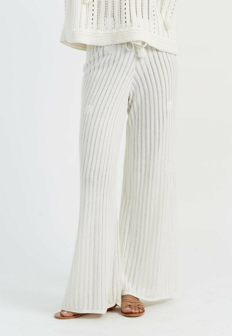 Pantaloni ampia in maglia bianca con strisce verticali e dettagli testurizzati, abbinati a sandali marroni con cinturini. Tessuto leggero.