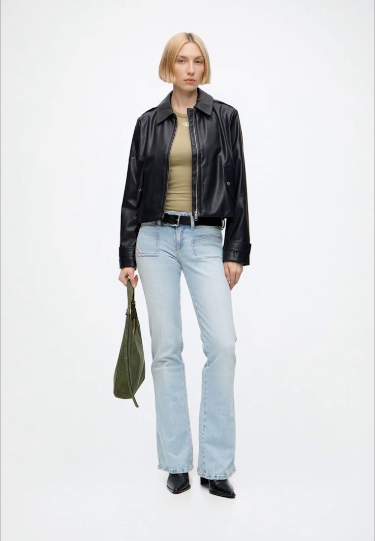Femme aux cheveux blonds courts, portant une veste en cuir noire, un haut vert olive, un jean bleu clair, des chaussures noires, tenant un sac à main vert.