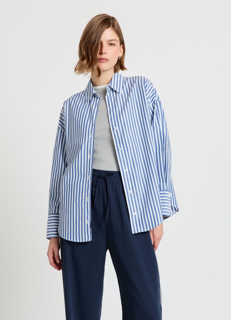 Chemise à rayures bleues et blanches avec col classique, manches longues et coupe décontractée, superposée sur un haut gris et un pantalon bleu marine.