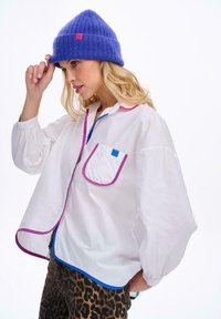 Bílá oversized košile s růžovým a modrým lemováním, s kapsou na levé straně hrudi. Fialová pletená čepice s růžovou nášivkou s logem. Kalhoty se zvířecím potiskem.