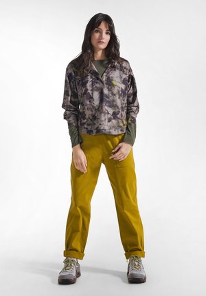 Une jeune femme se tient face à l’avant, portant un haut à manches longues à motif floral, un pantalon jaune moutarde et des baskets grises.