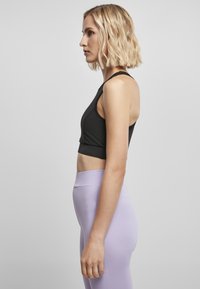 Črn skrajšan atletski top s križnim hrbtnim delom, v kombinaciji z visokimi svetlo vijoličnimi leggins hlačami. Gladek material in prilegajoča silhueta.
