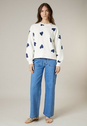 Femme portant un sweat-shirt blanc avec des motifs de cœurs bleus marine, un jean bleu droit et des sandales marron clair, debout devant un fond uni.