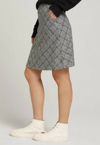 Mujer vestida con una falda cuadros gris con bolsillos, un suéter negro y zapatillas altas blancas, de pie frente a un fondo liso.