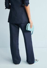 Blazer marine à manches retroussées et accents de boutons, associé à un pantalon à jambes larges. Tenant un sac à main texturé bleu clair. Sandales avec des lanières.