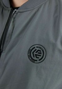 Chaqueta bomber gris con un logo circular negro en el pecho y detalles texturizados. Cierre con cremallera en el frente y cuello acanalado.